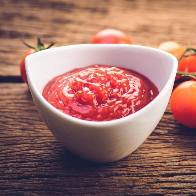 Yoğun kıvamlı ve parlak kırmızı renkteki ev yapımı domates sosu, cam bir kase içerisinde, üzerine serpiştirilmiş taze fesleğen yapraklarıyla doğal ve iştah açıcı bir şekilde sunuluyor.