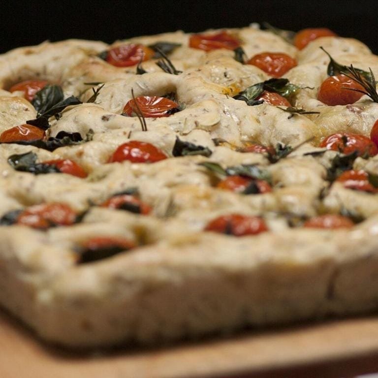 Fırından yeni çıkmış, domates ve biberiyeyle süslenmiş nefis bir focaccia ekmeği, üstü altın rengi kızarmış ve buhusu hala üstünde duruyor.