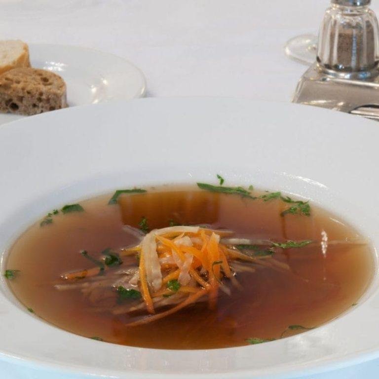 Konsome Çorbası Tarifi: Şık bir porselen kasede, kristal berraklığında ve altın rengiyle parlayan, taze otlarla süslenmiş, yoğun lezzetli ve rafine kıvamlı konsome çorbası.
