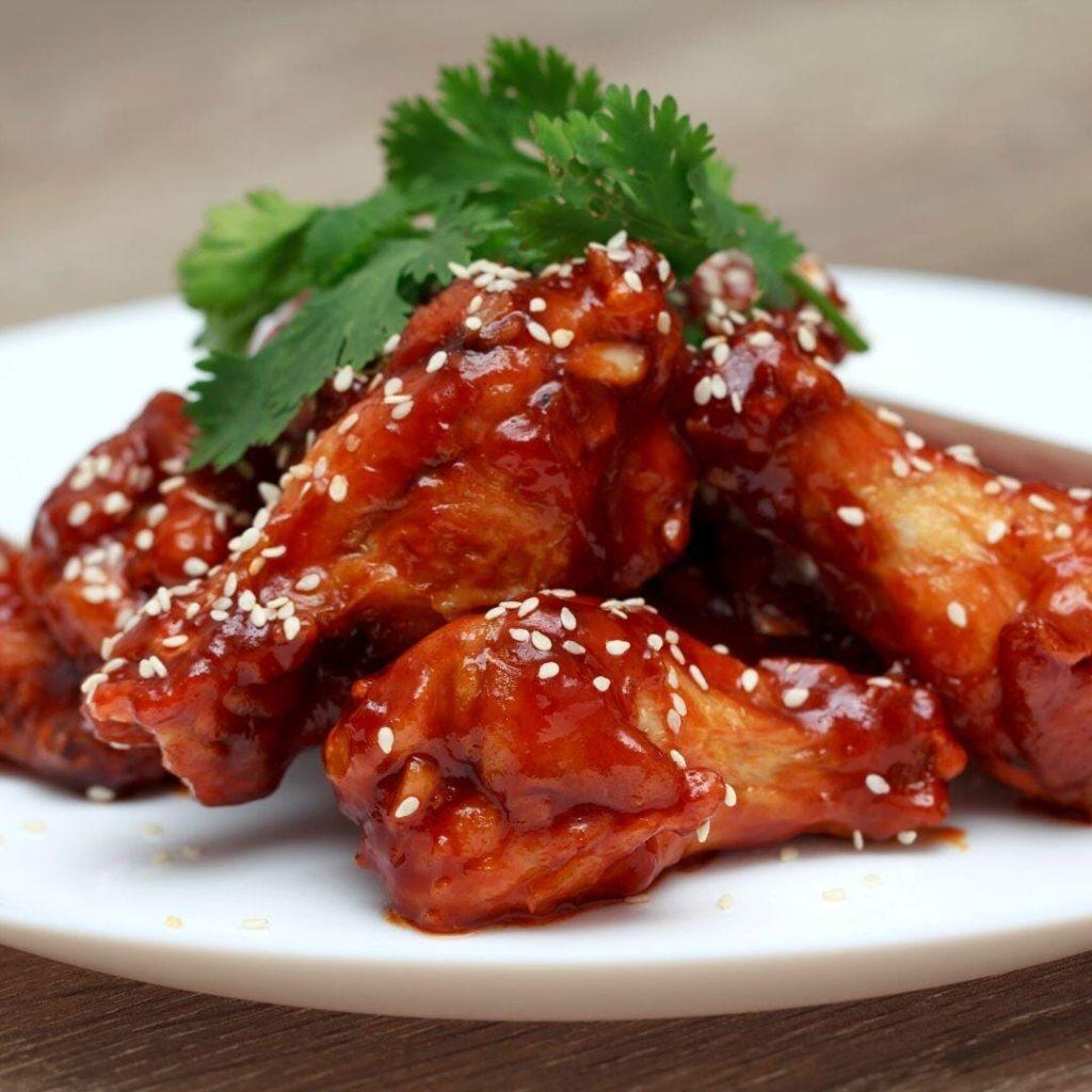Chimaek Tarifi (치맥) Kore usulü Chimaek tarifi: Yapışkan gochujang sosu ile kaplanmış, üzerinde kavrulmuş susam taneleri olan çıtır tavuk kanatları servis tabağında sunuluyor.