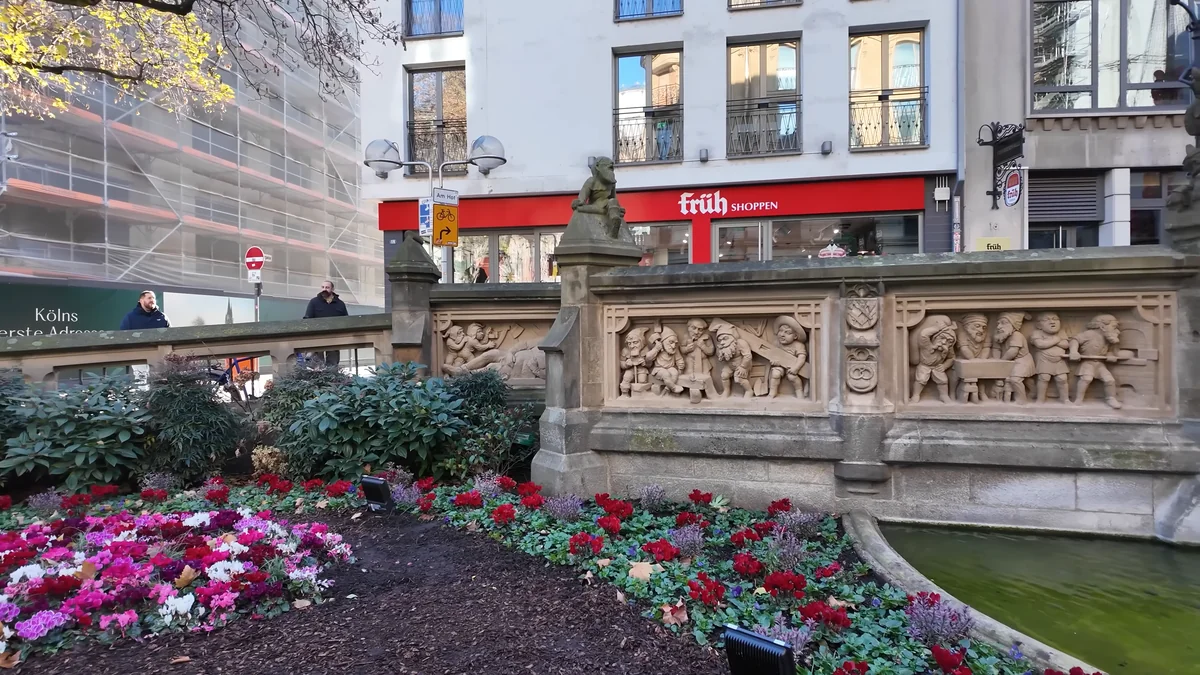 The carved stone wall of the Heinzelmännchen fountain in Cologne with flowers in front