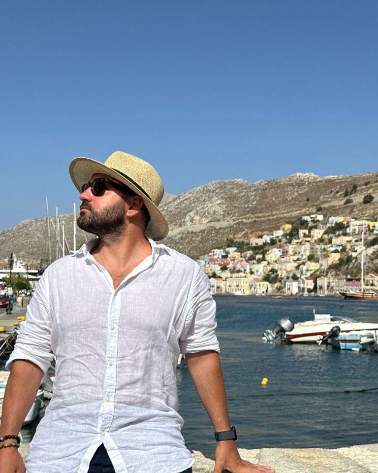 Symi Adası sahilinde beyaz gömlekli bir turist ve arka planda adanın meşhur renkli evlerinin manzarası.