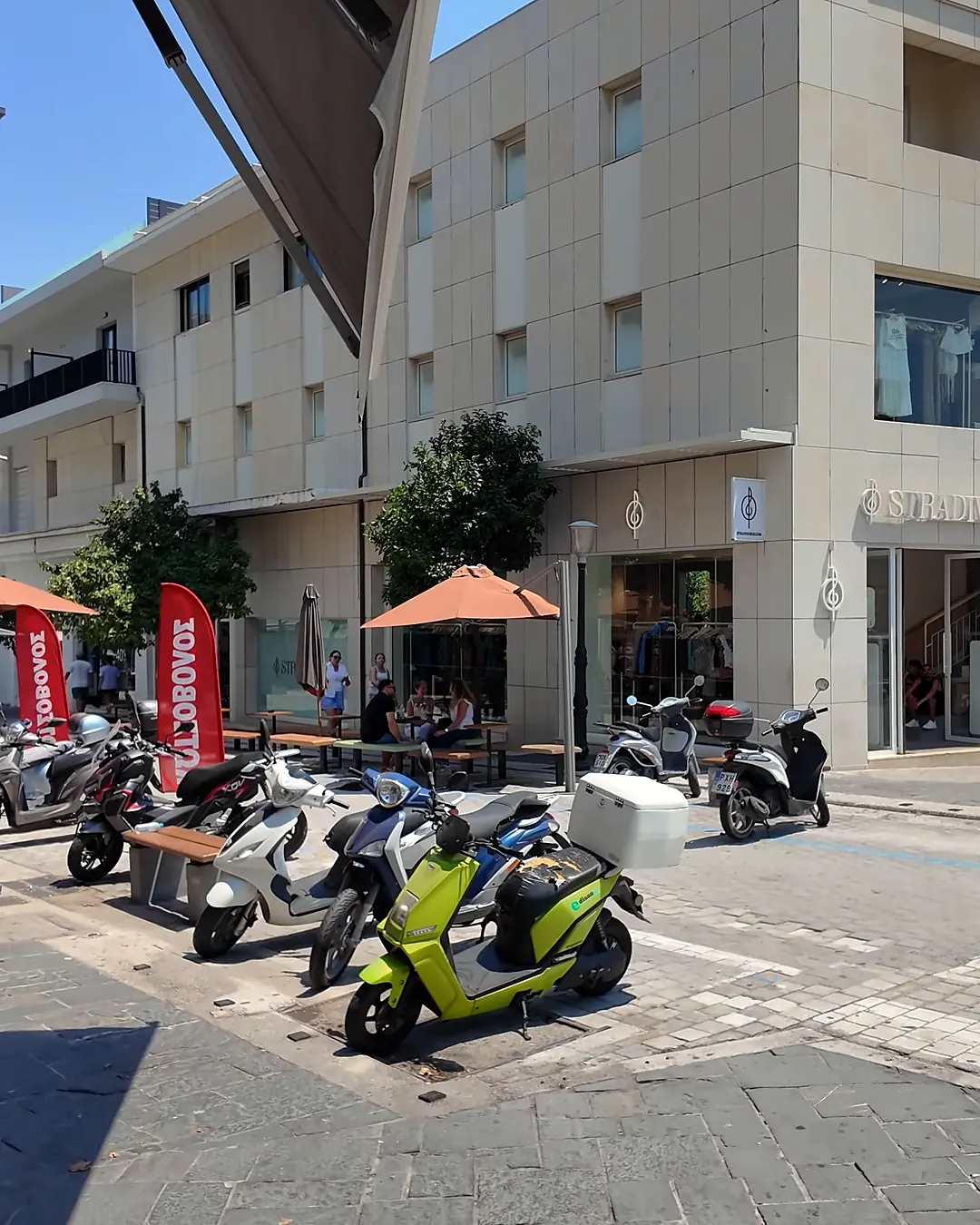 Rodos alışveriş sokağında Stradivarius mağazası, gölgelikli kafe masaları ve kaldırıma park edilmiş scooterlar