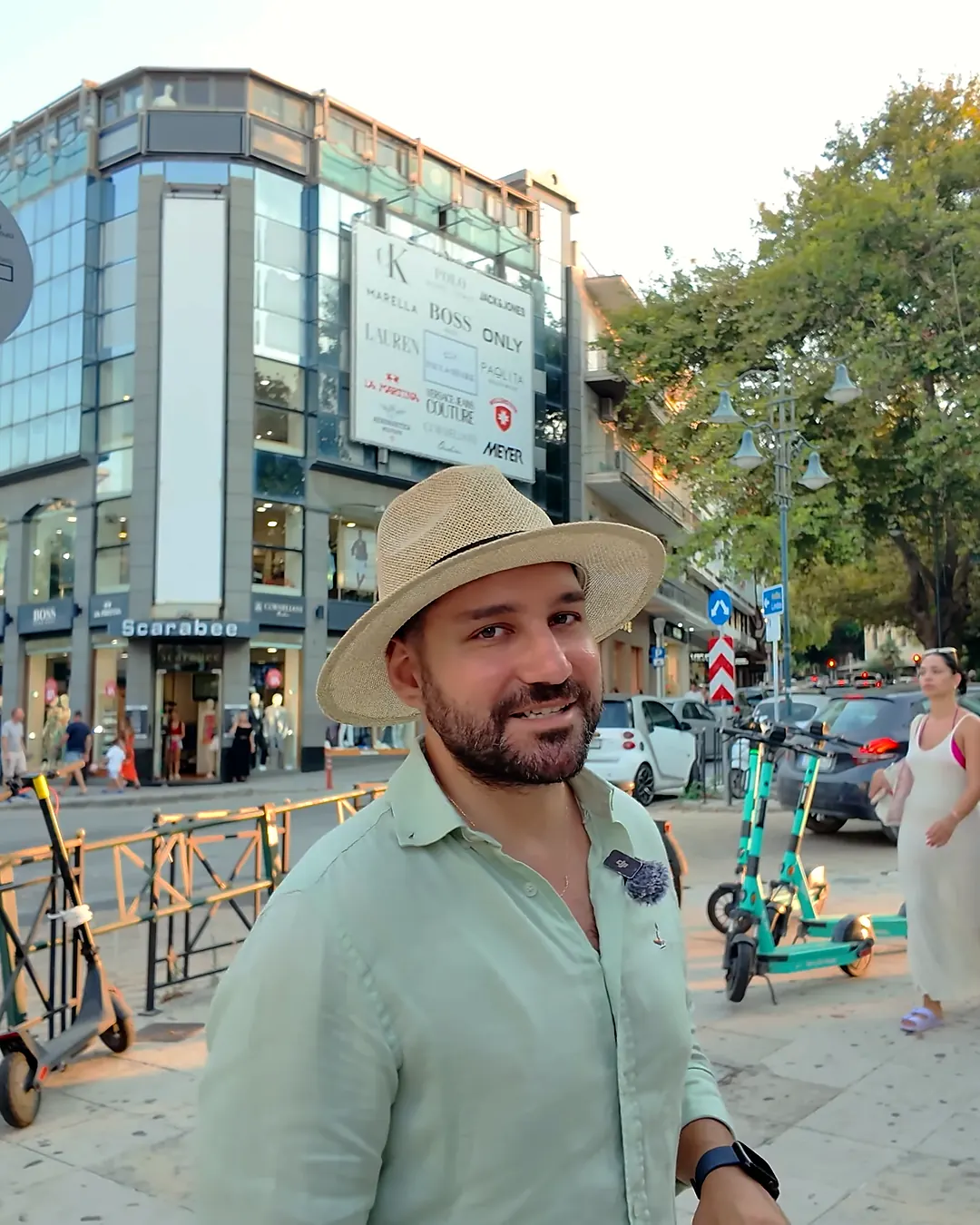Rodos yeni şehirdeki alışveriş caddesinde markalı mağaza vitrinleri, elektrikli scooterlar ve akşamüstü yürüyen kalabalık