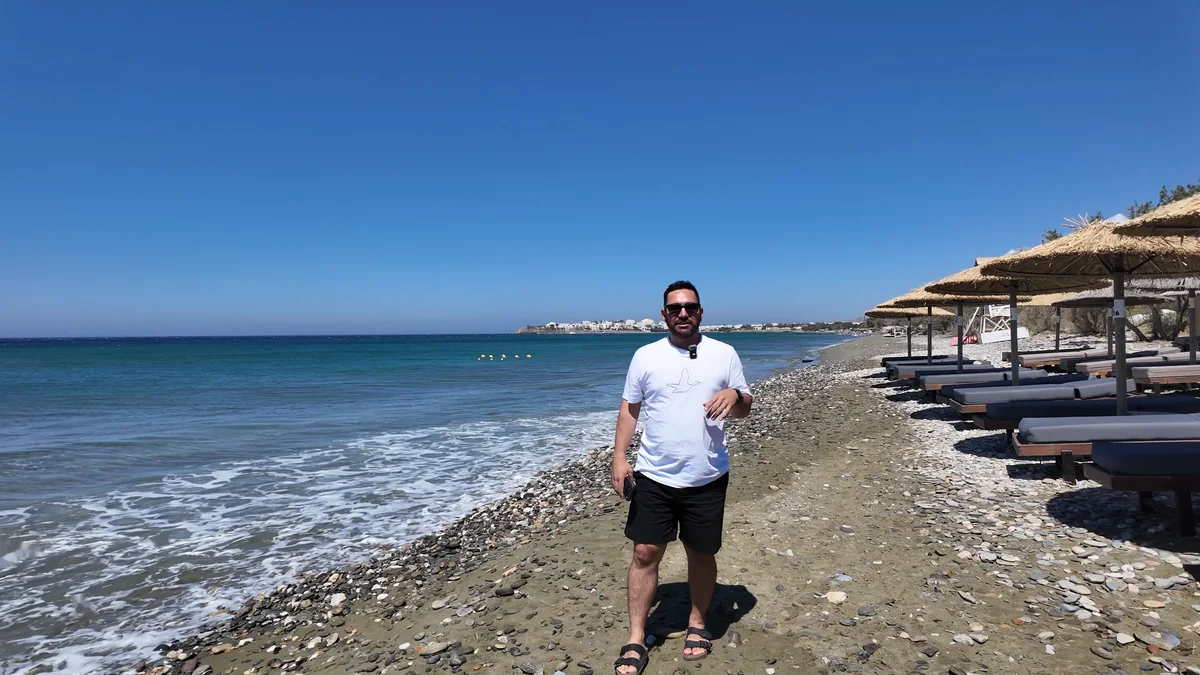 Tinos'ta Agiafos Plajı: çakıl taşlı sahil, hasır şemsiyeler, şezlonglar ve uzakta ada merkezi