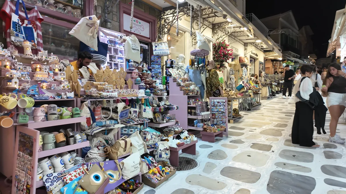 Tinos Çarşısı gece: hediyelik eşya dükkanları, seramikler ve mermer döşeli caddede gezen insanlar