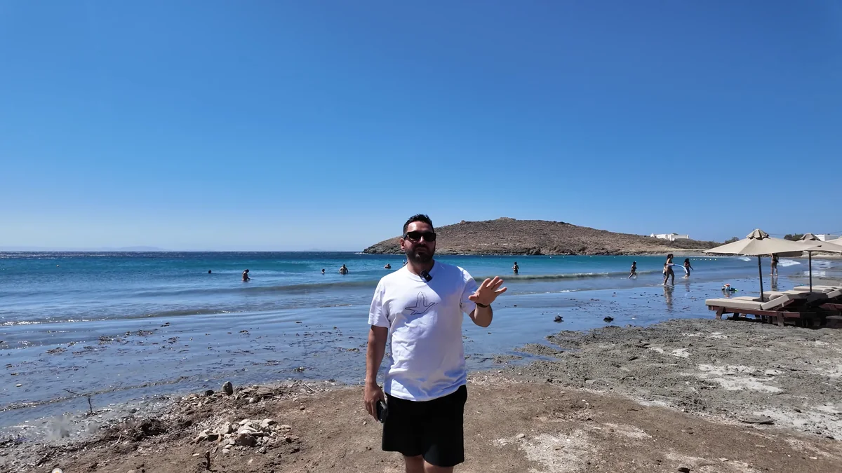 Tinos'ta Inas Plajı: kumlu sahil, arkada kayalık burun, denizde yüzenler ve sahilde şezlonglar