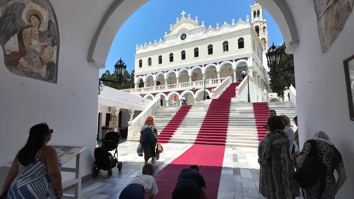 Tinos'ta Panagia Evangelistria Kilisesi: kemerli girişten görünen cephe ve merdivenlere serilmiş kırmızı halı