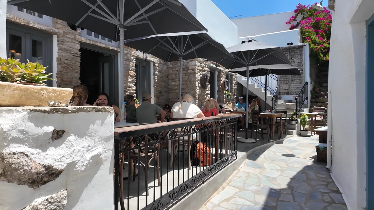 Tinos’ta Volax köyündeki Kafenai: taş duvarlı sokakta şemsiyeli teras ve oturan misafirler