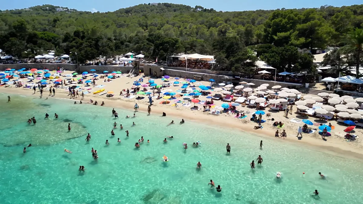 İbiza’da Cala Bassa plajı: geniş kumsal, renkli şemsiyeler ve sığ turkuaz deniz