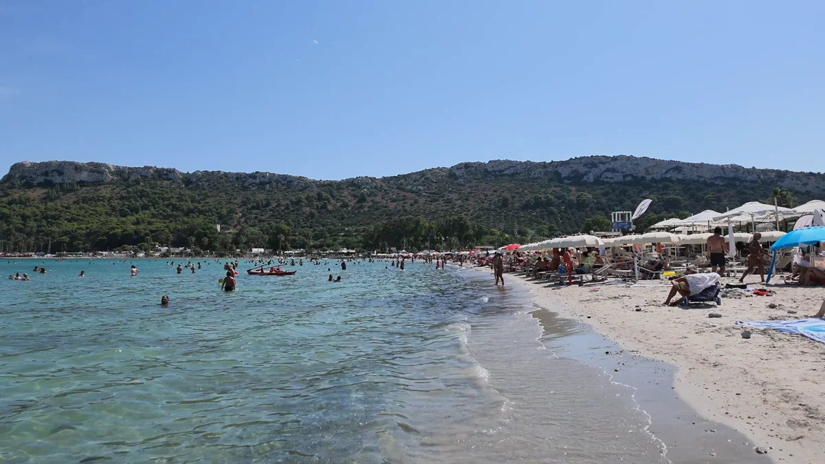 Cagliari Poetto plajında sığ berrak deniz, şezlonglar ve arkadaki yeşil tepeler