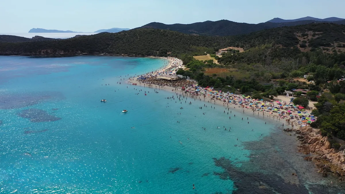 Teulada Tuerredda plajı: turkuaz koy, kumsaldaki şemsiyeler ve arkadaki yeşil tepeler